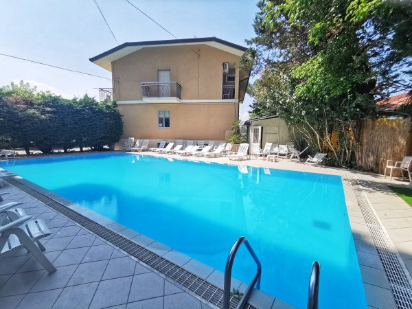 Piscina azzurra all'aperto con lettini bianchi e una casa sullo sfondo.