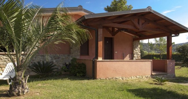 Esterno di una casa con veranda coperta, pareti rosa e prato con palme.