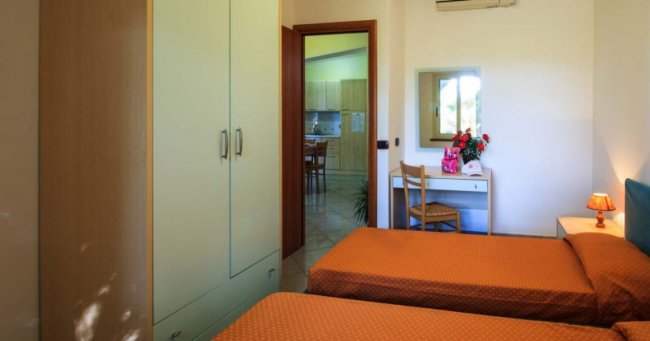 Camera con due letti singoli, armadio bianco e scrivania. Porta aperta verso la cucina.