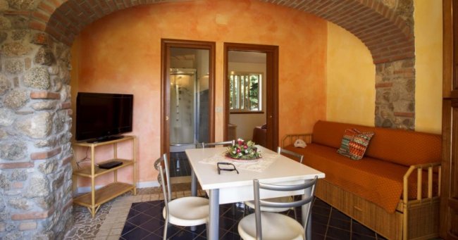 Sala con tavolo bianco, divano arancione e muri in pietra. Due porte sullo sfondo.