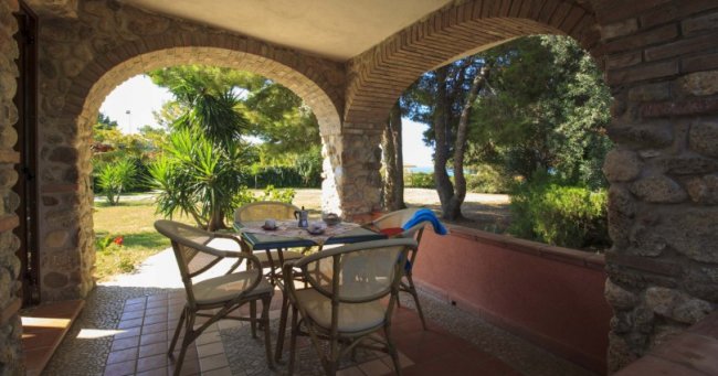 Veranda con tavolo e sedie sotto archi in pietra, circondata da alberi e vegetazione.