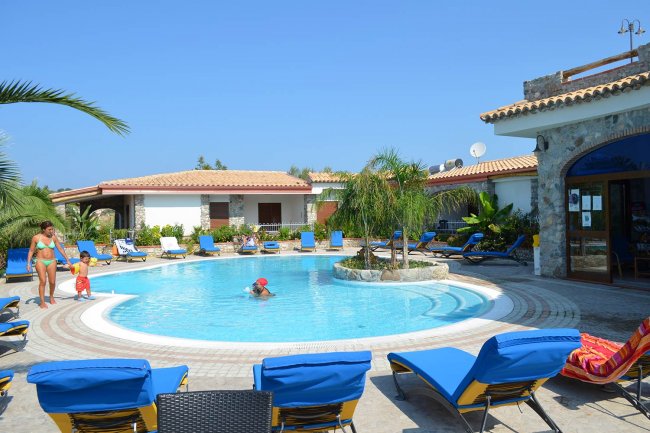 Piscina circolare con lettini blu, circondata da casette e palme sotto un cielo azzurro.