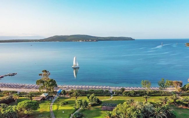Barca a vela sul mare con spiaggia e colline sullo sfondo.