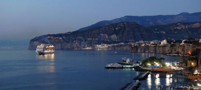 Vista notturna su un porto con una nave e montagne in lontananza.