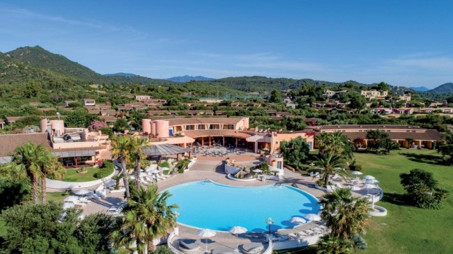 Vista aerea di un resort con piscina circondata da palme e ombrelloni bianchi.